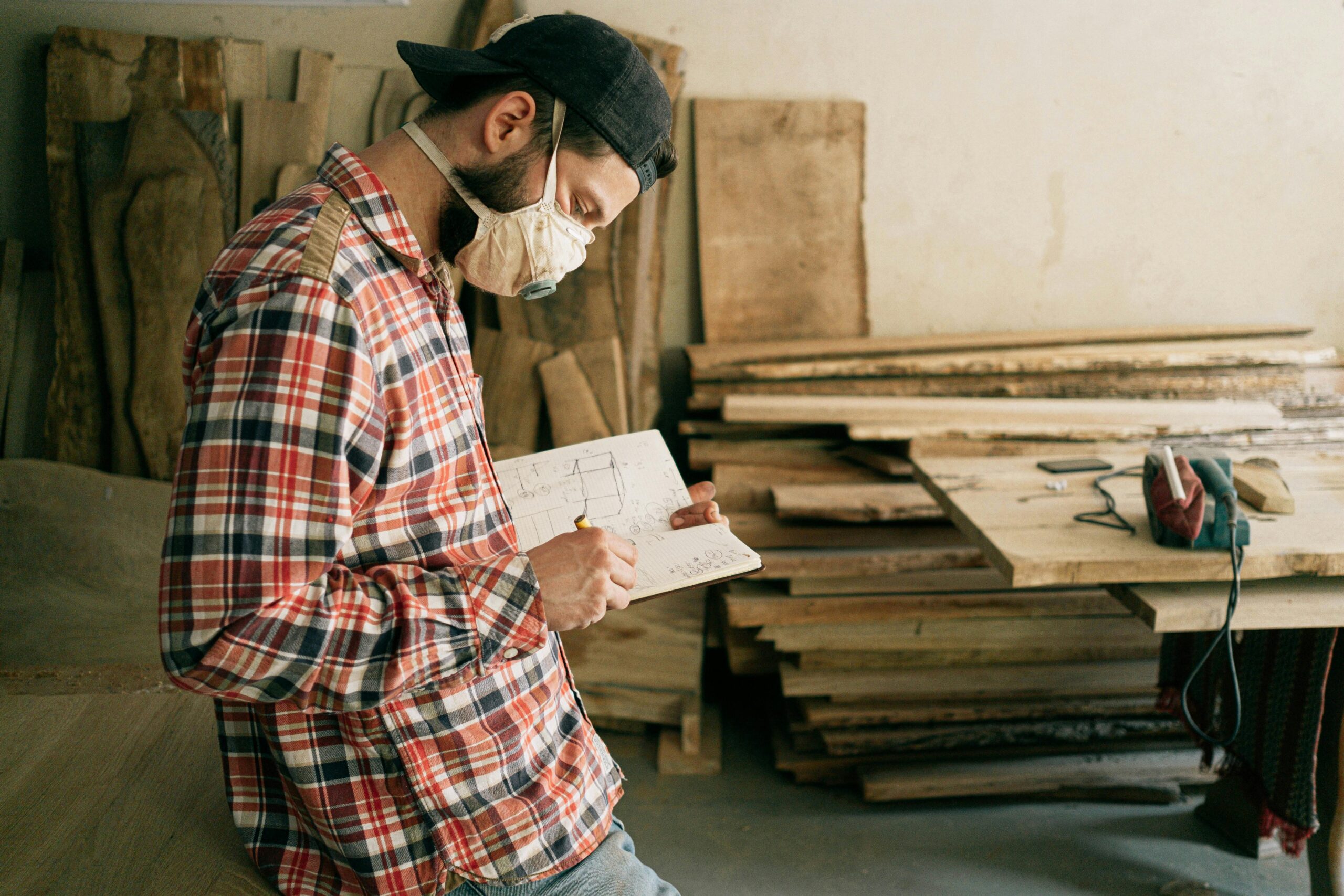 A craftsman wearing a mask sketches plans in a woodwork studio.